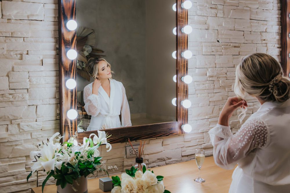 A woman in white robe adjusts her elegant updo hairstyle while looking in a mirror with vanity lights.
