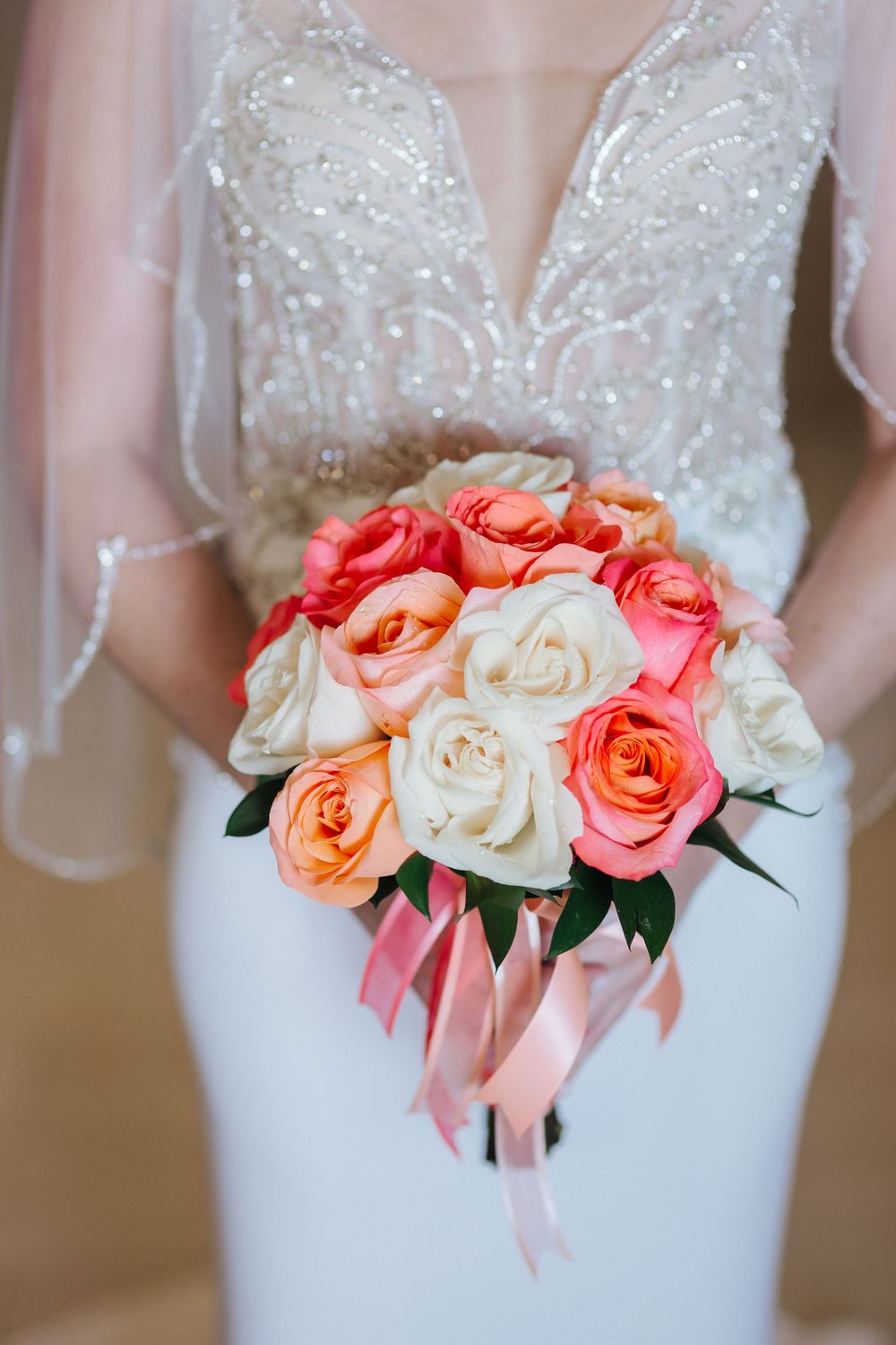 Beautiful bridal bouquet featuring coral and white roses with pink ribbons against an elegant beaded wedding dress.