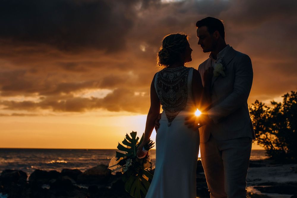 Silhouette of newlywed couple standing against dramatic orange sunset sky at beach wedding.