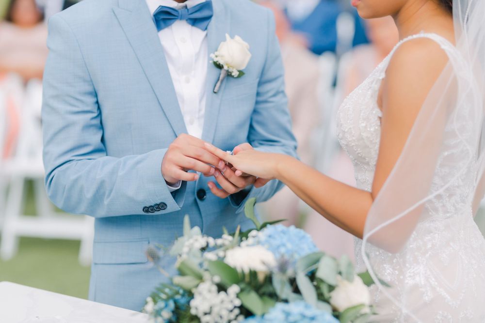 Close-up shot of exchanging rings during outdoor wedding ceremony with light blue suit and white floral bouquet.