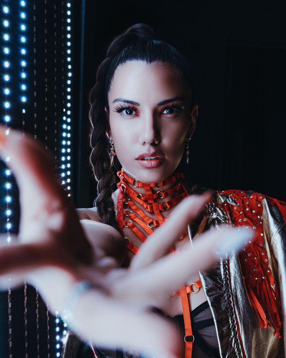 Dramatic portrait with neon lights showing person in orange high neck top reaching toward camera.