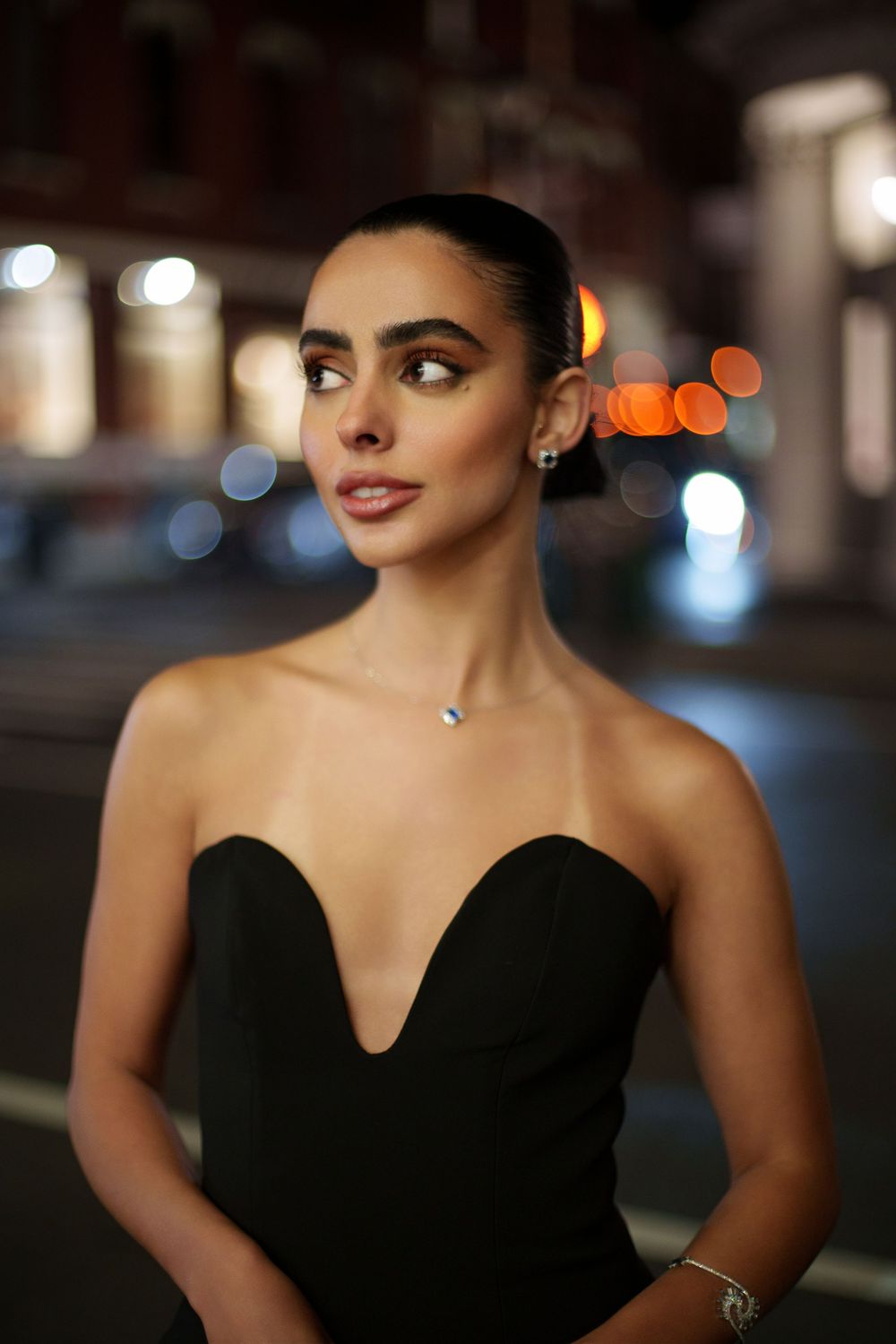Portrait of someone in a black strapless sweetheart neckline dress at night with city lights in background.