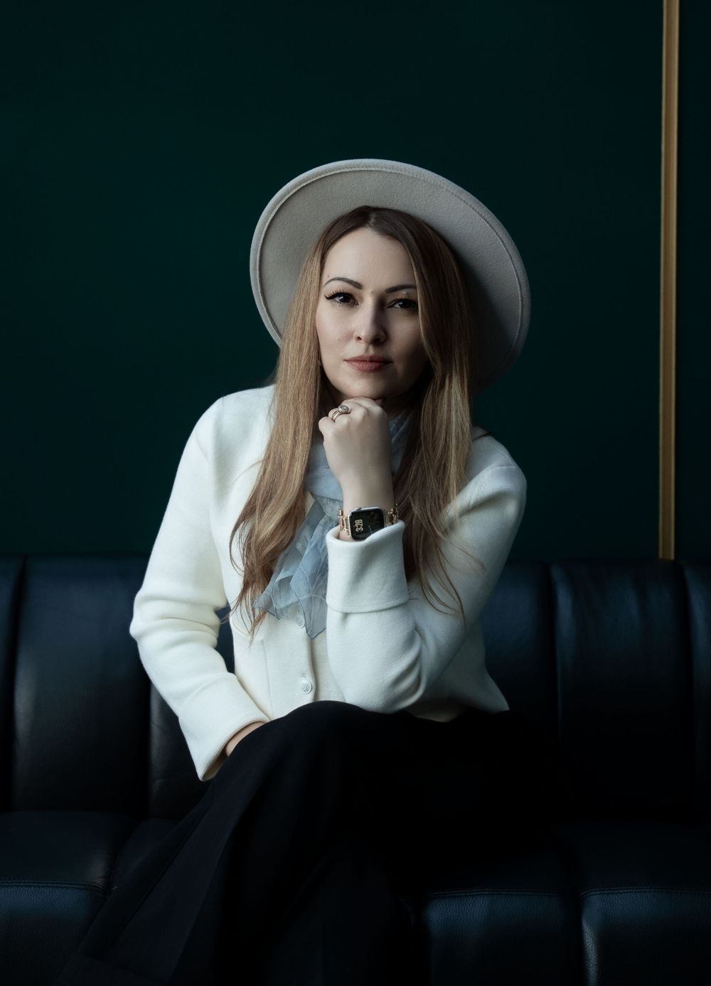 Fashion portrait of person in white sweater and hat against dark background lounging on leather couch.