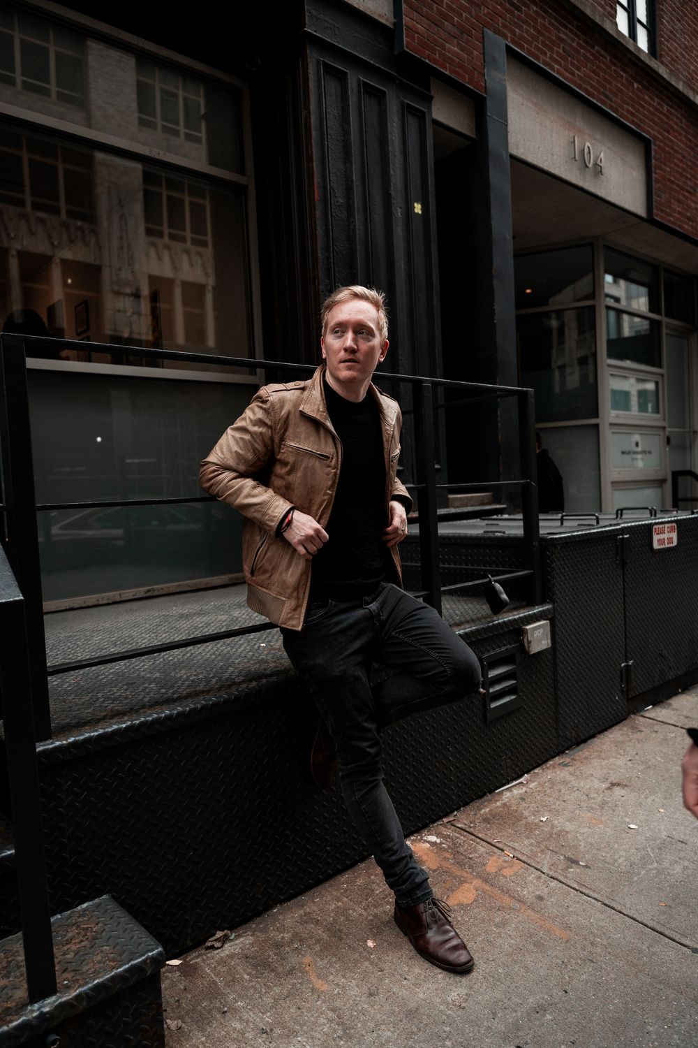 A person in a brown leather jacket and black pants poses on steps outside an urban brick building.