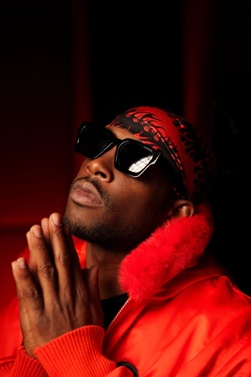 Dramatic portrait of a person in sunglasses and red clothing against dark background with hands clasped in prayer pose.