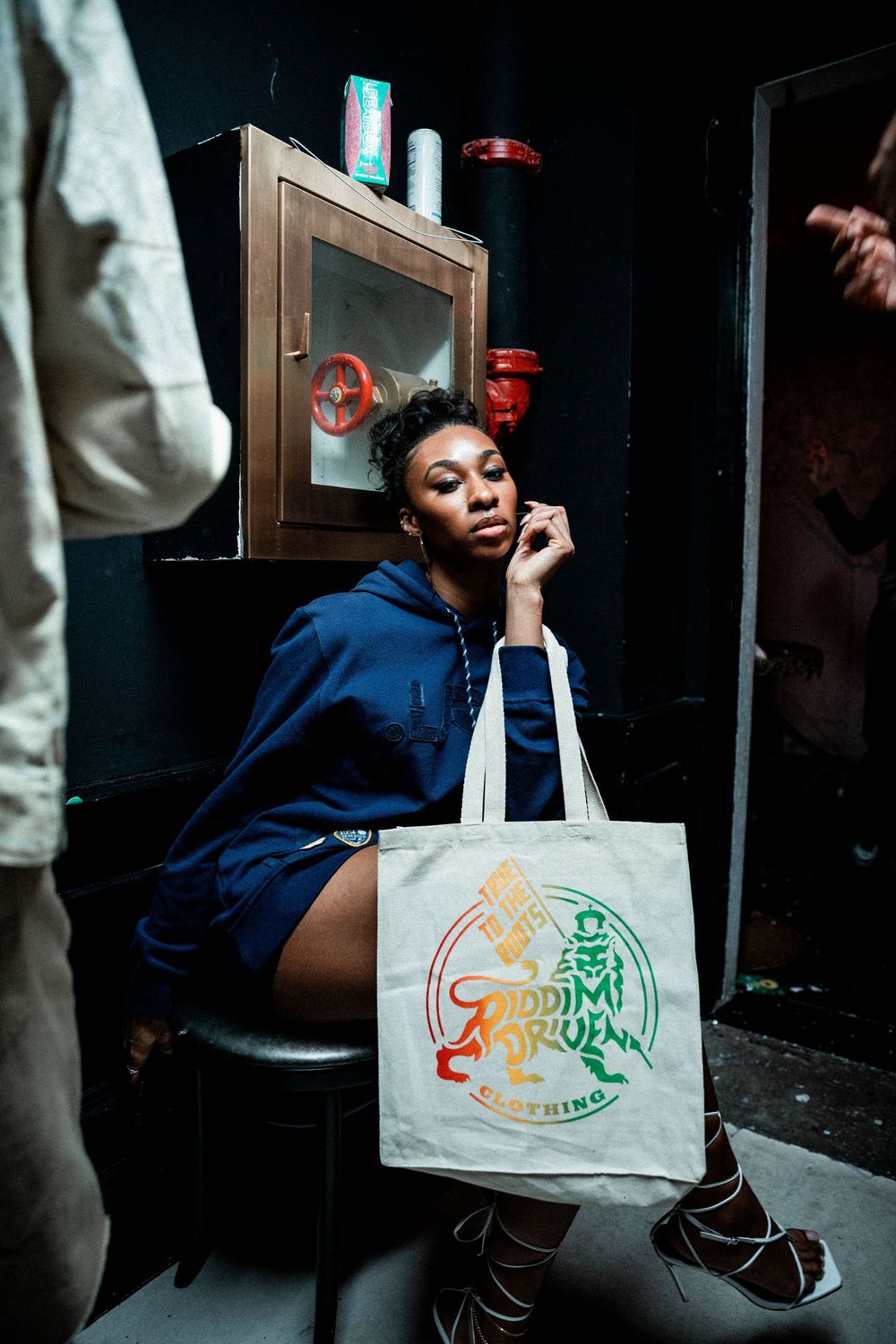 A person wearing a blue dress and holding a tote bag sits in a dark interior setting.