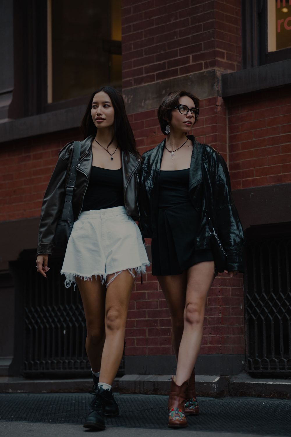Two people in leather jackets and contrasting outfits pose against brick building.