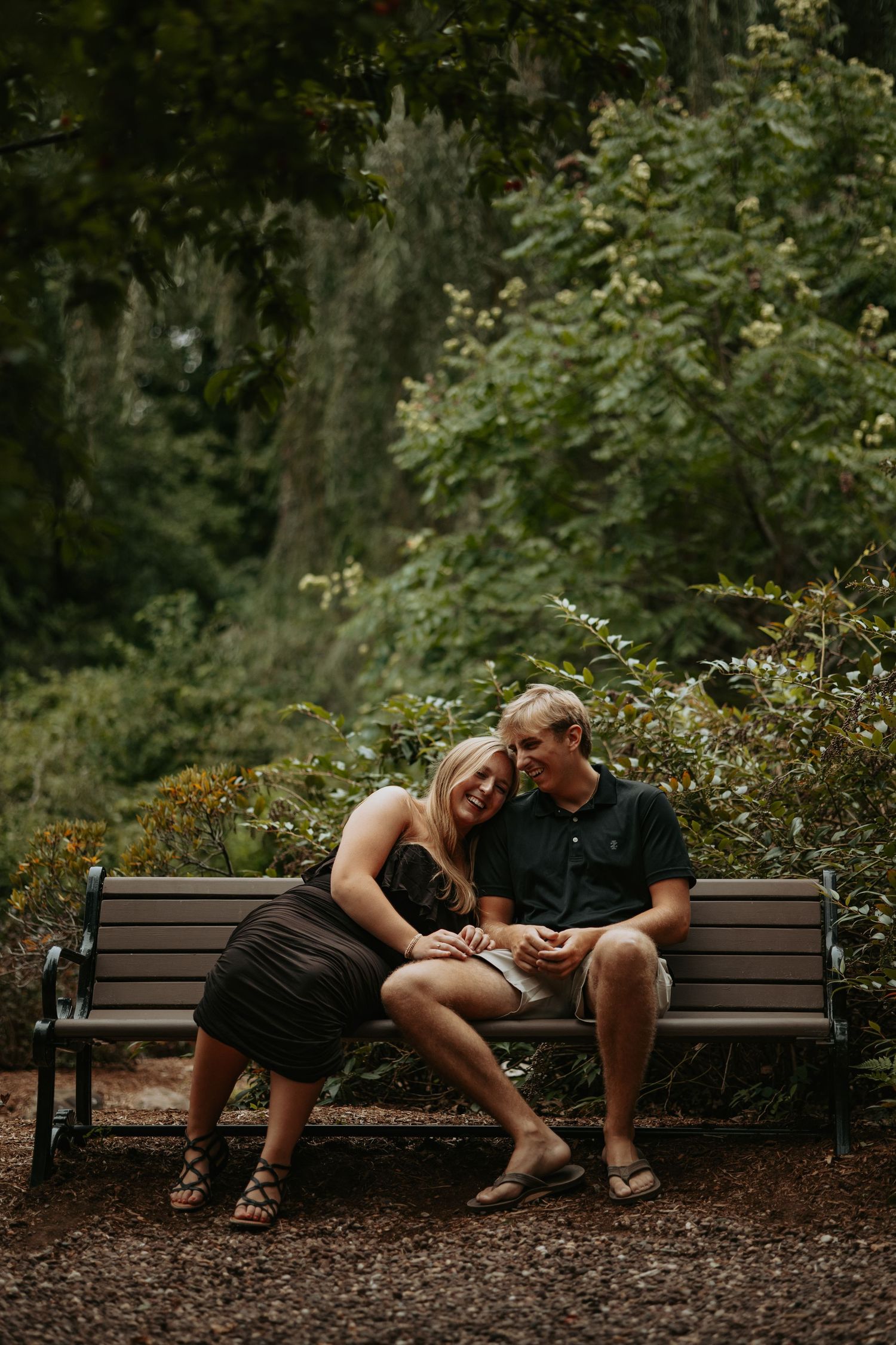 Smiling couple sits close together on a wooden park bench, the woman leaning her head on the man's shoulder, surrounded by lush green trees.