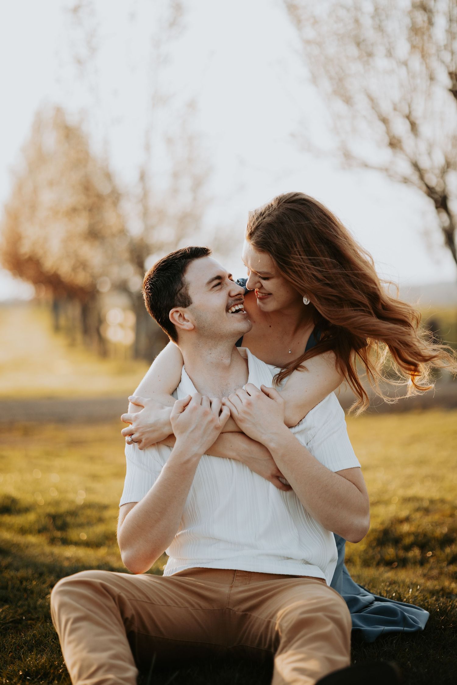 A happy couple embraces outdoors at golden hour, the woman hugging the man from behind as they smile at each other.