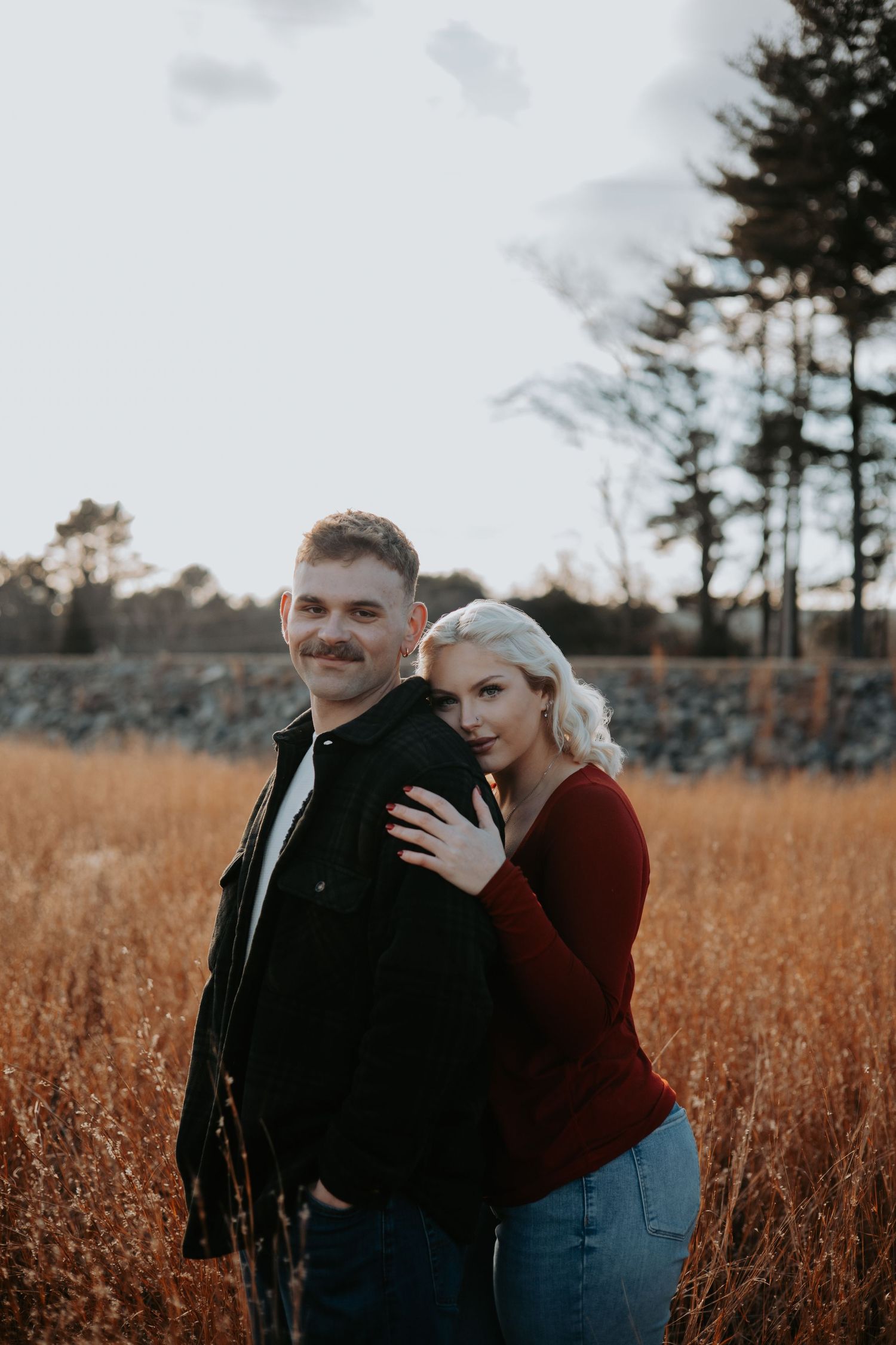 Couple embraces in a golden autumn field at sunset with evergreen trees in background at Mansfield Hollow State Park.