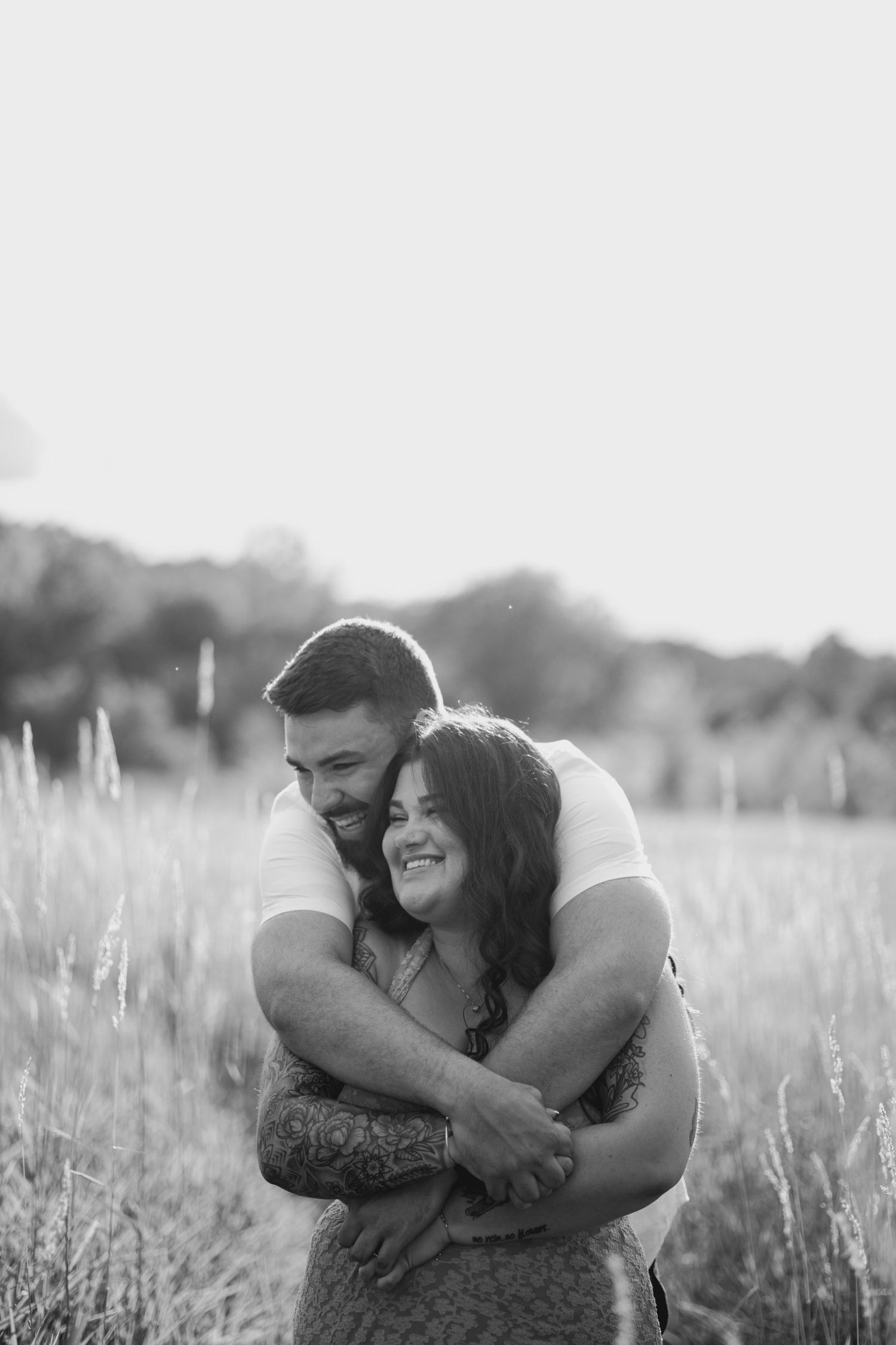 Black and white photo of a smiling couple embracing in a tall grass field, the man hugging the woman from behind at sunset.