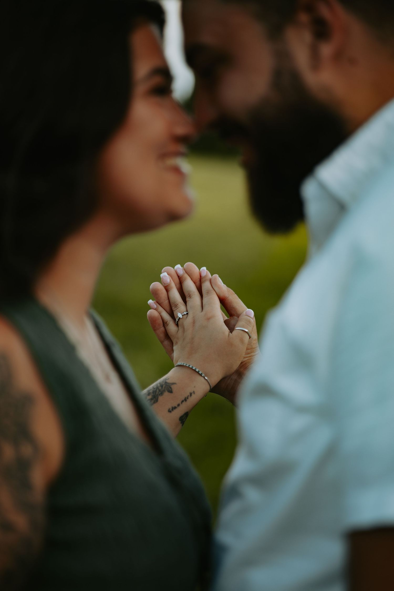 Smiling couple standing face to face with clasped hands and rings in focus, outdoors on a blurred green lawn.