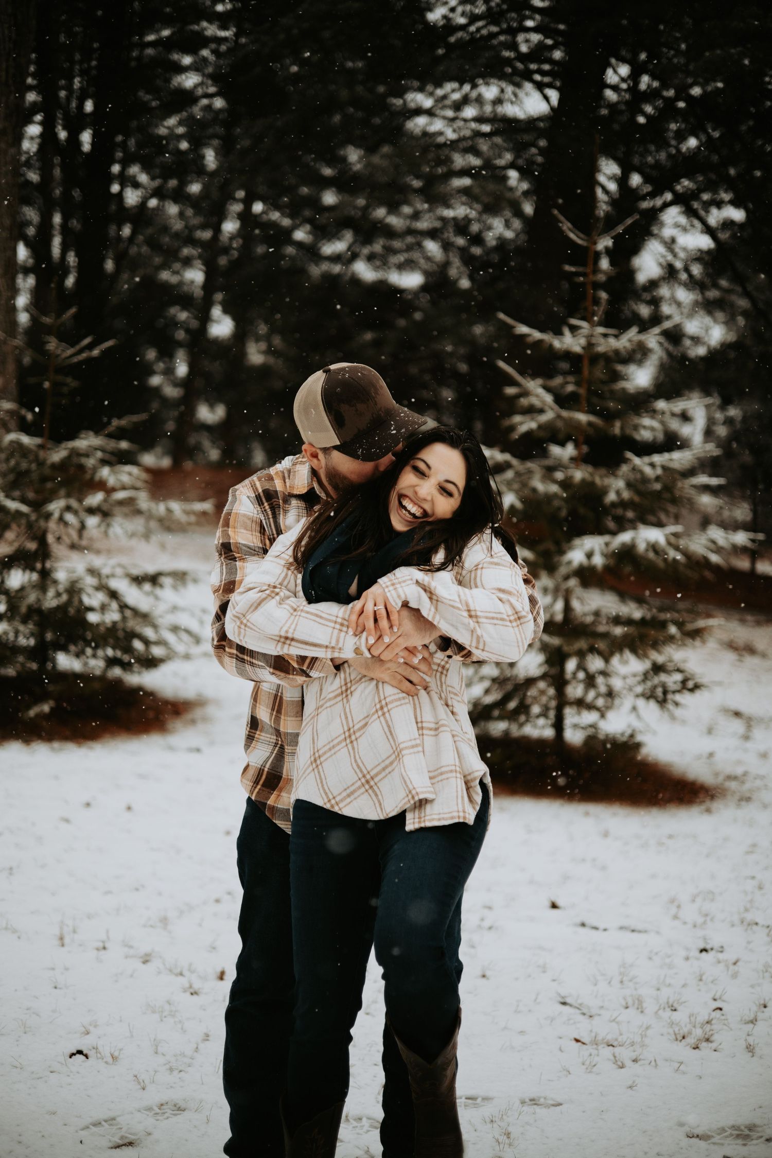 Couple embracing in snowy winter forest, woman smiling as man hugs her from behind, both wearing plaid flannel shirts.