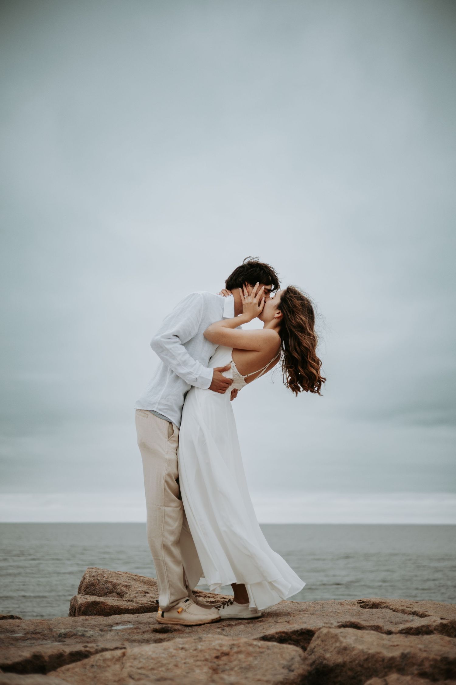 Couple sharing a romantic kiss on rocky shoreline, woman in white dress, man in casual attire, overcast ocean backdrop.