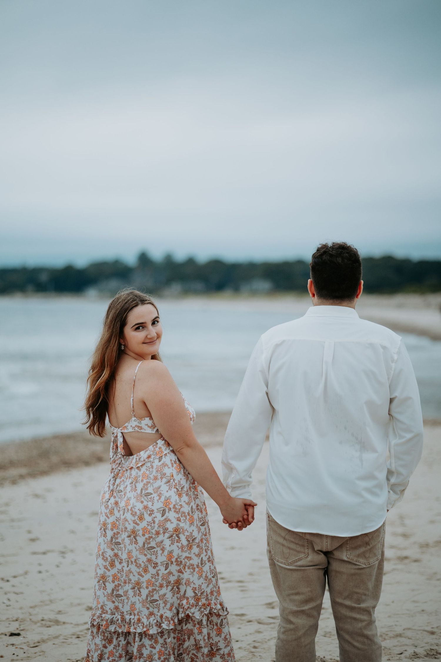 Smiling woman in a floral sundress holds hands with a man in a white shirt while walking along a calm, overcast beach.