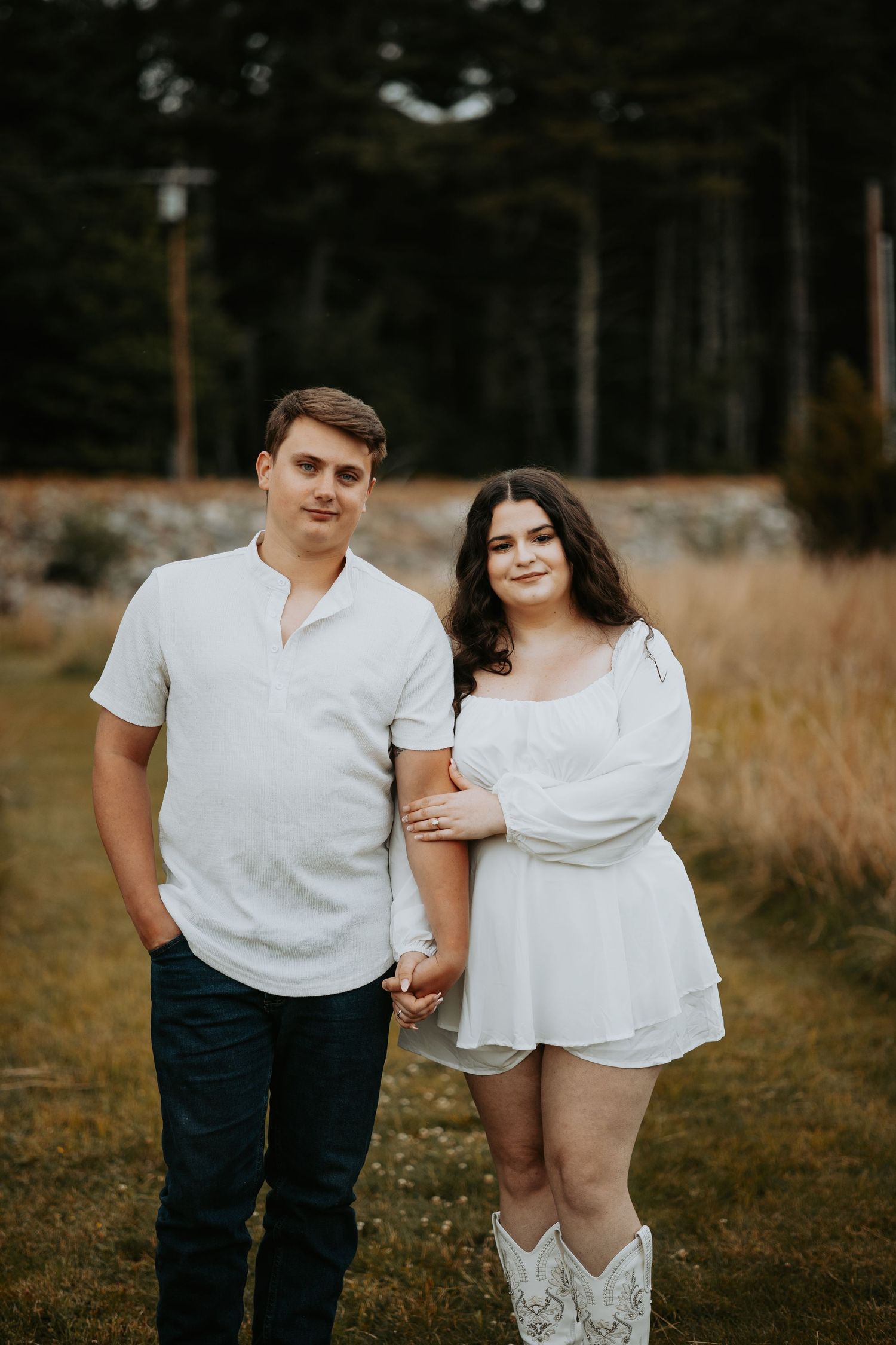 A smiling couple holds hands while posing outdoors in a grassy field, she in a white dress and cowboy boots, he in a white shirt and jeans.