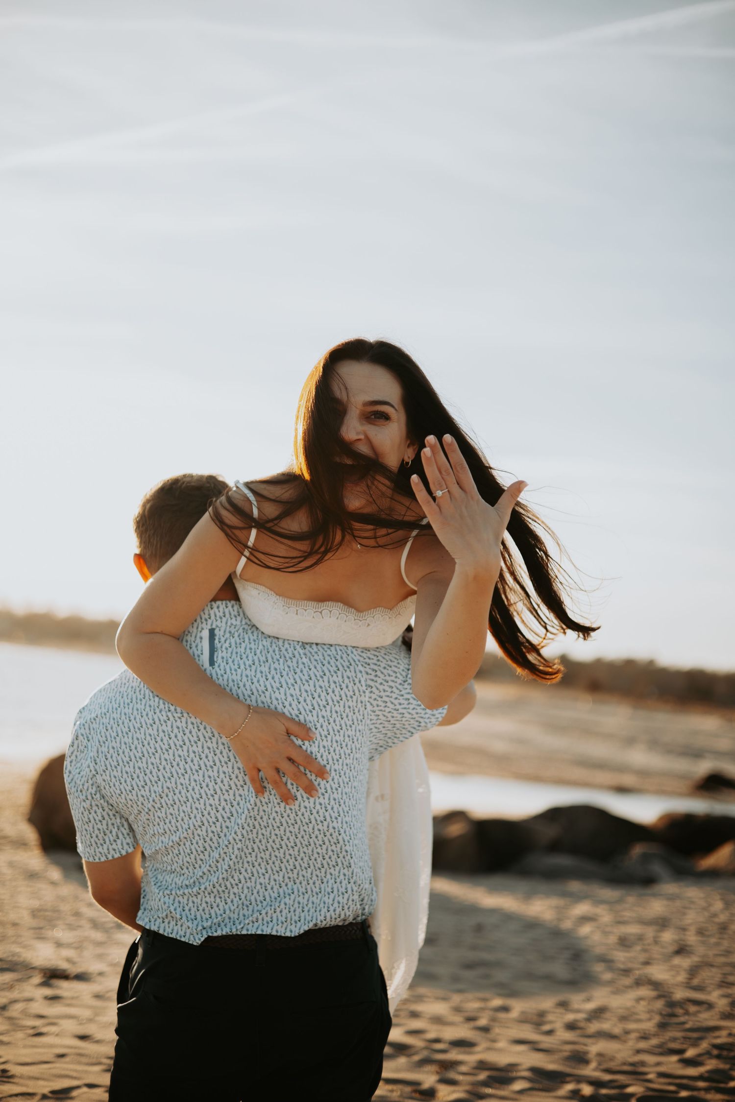 Couple sharing a romantic piggyback ride on a sandy beach at golden hour, woman showing engagement ring.