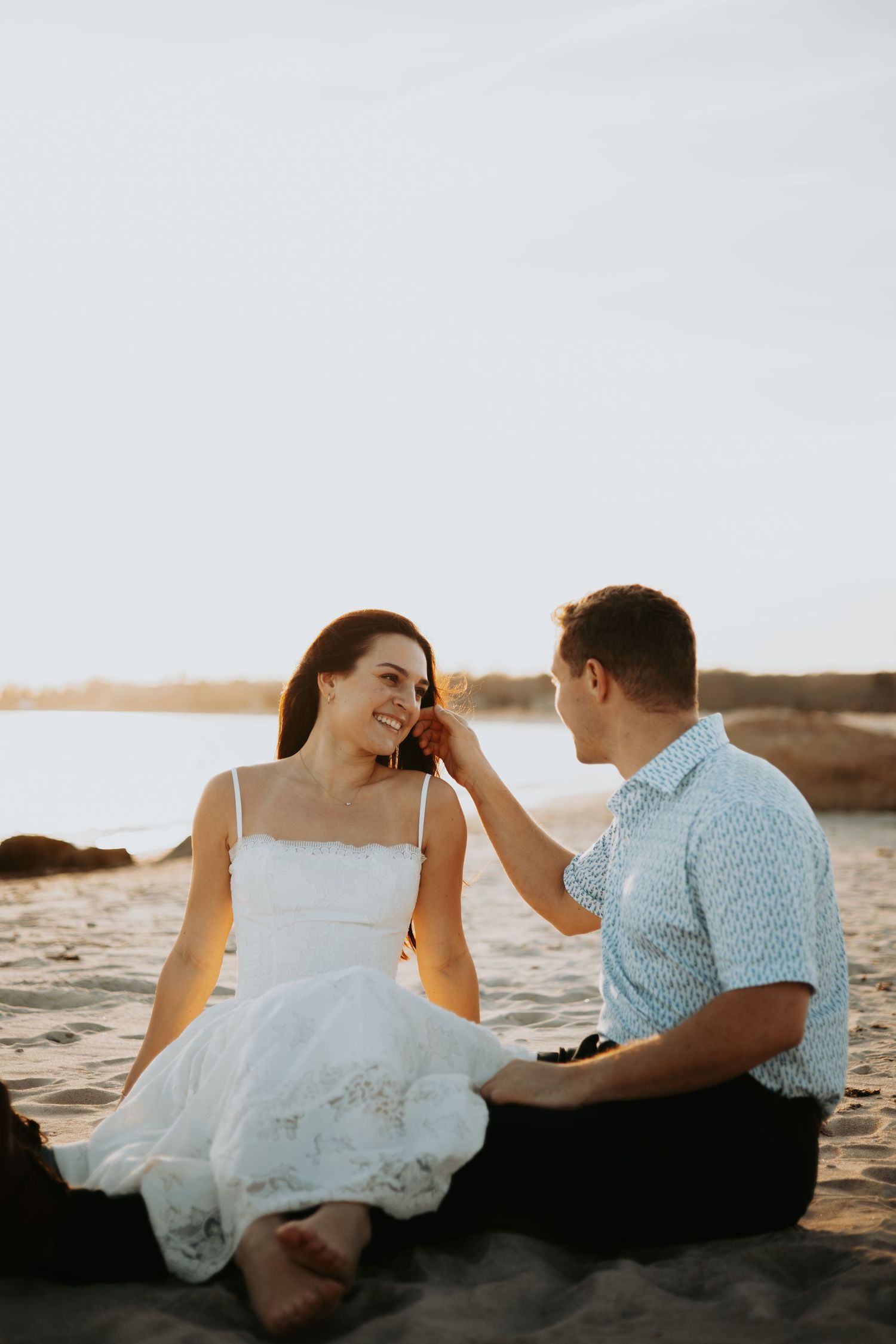 A couple sits on a sandy beach at sunset, sharing a tender moment as he gently touches her hair.