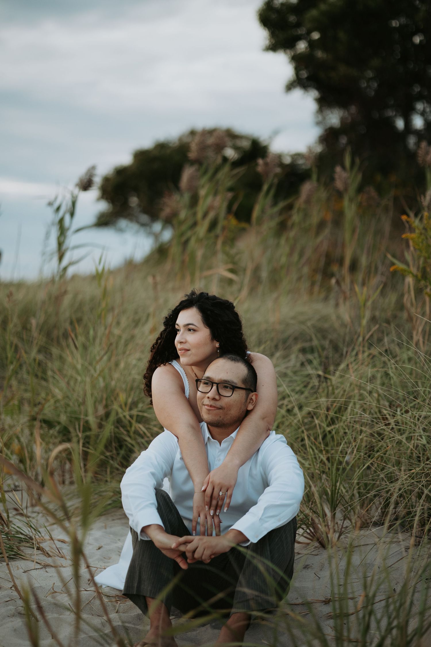 A couple sits close on a sandy beach dune as a woman hugs a smiling man in glasses from behind, surrounded by tall grass and trees.