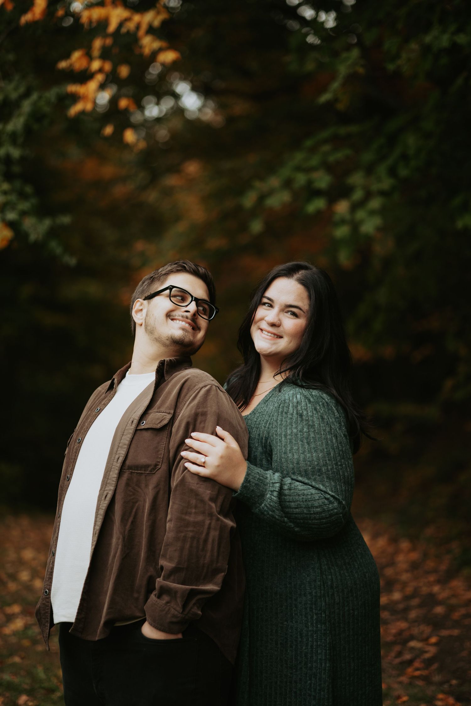 Smiling couple in cozy fall outfits embracing outdoors among autumn trees and fallen leaves during an engagement photo session.
