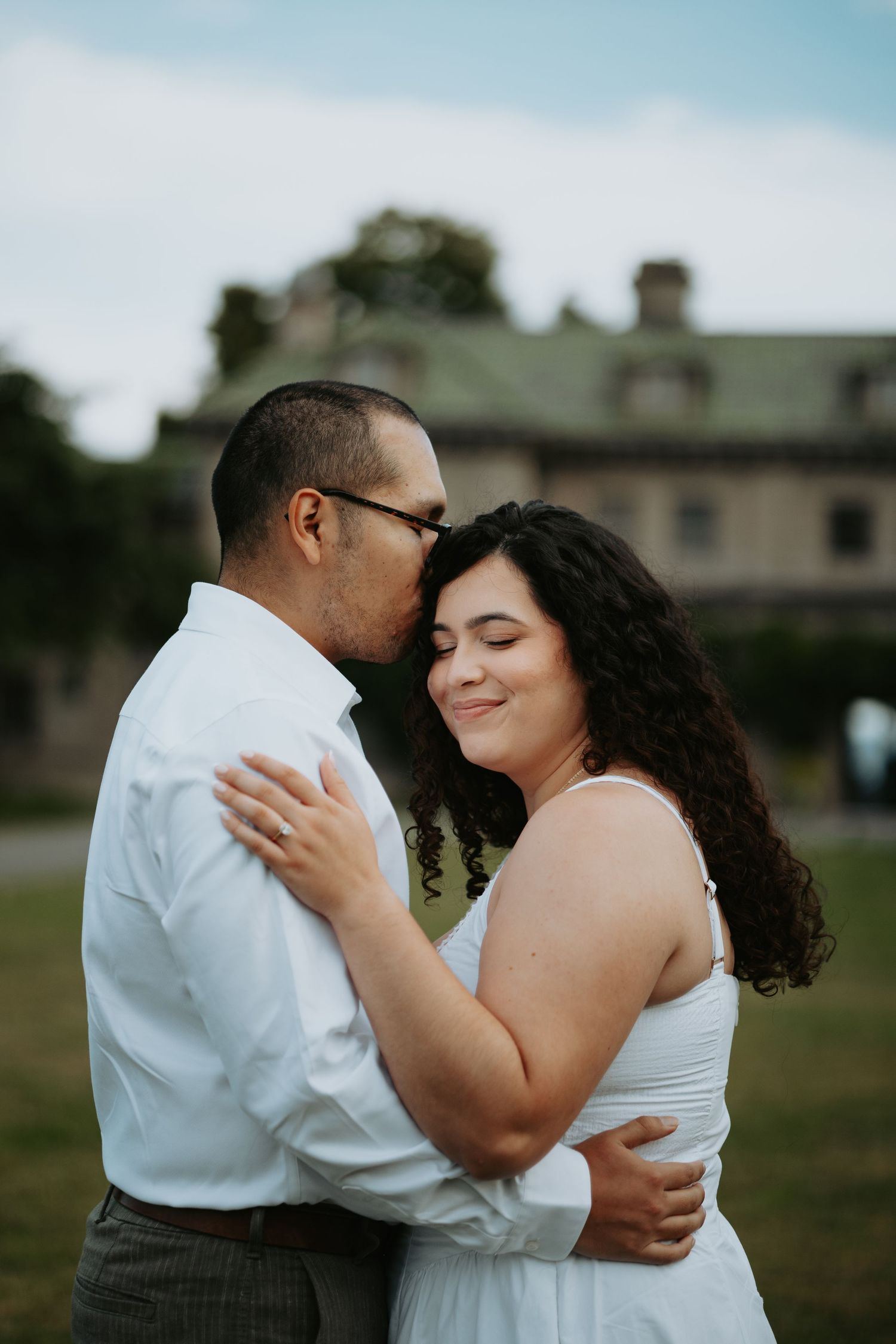 Man kissing the forehead of a smiling woman in a white dress during an outdoor engagement photo session with a historic mansion behind them.