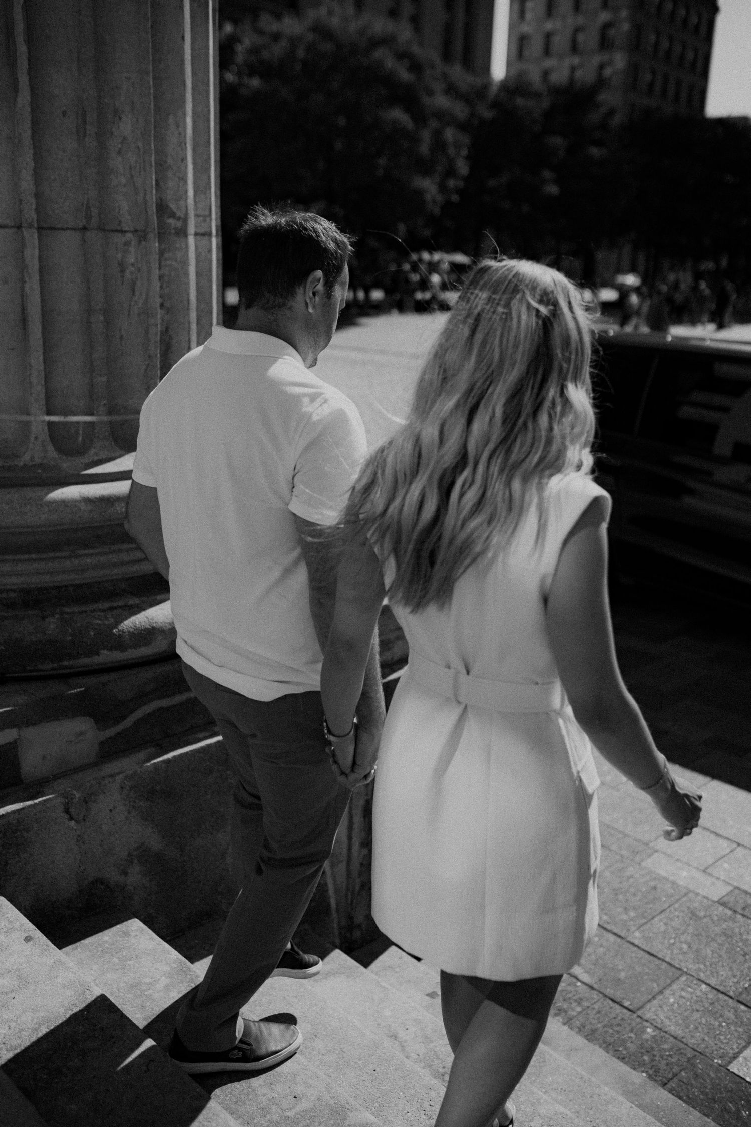 Black and white photo of a couple holding hands walking up stone steps near a classical building column.