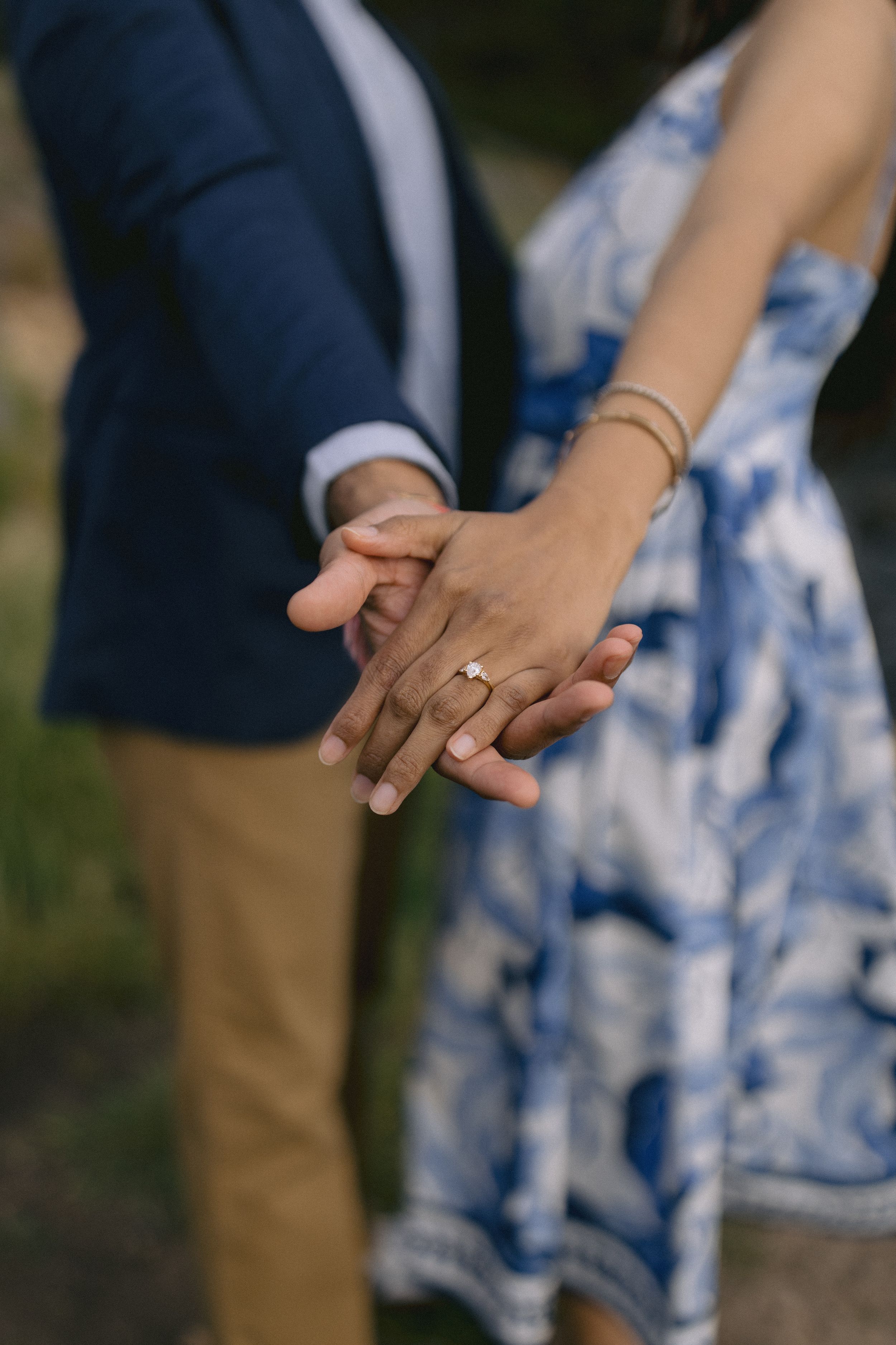 Close-up of couple holding hands and wearing rings, with blue floral dress and navy blazer visible against outdoor setting.