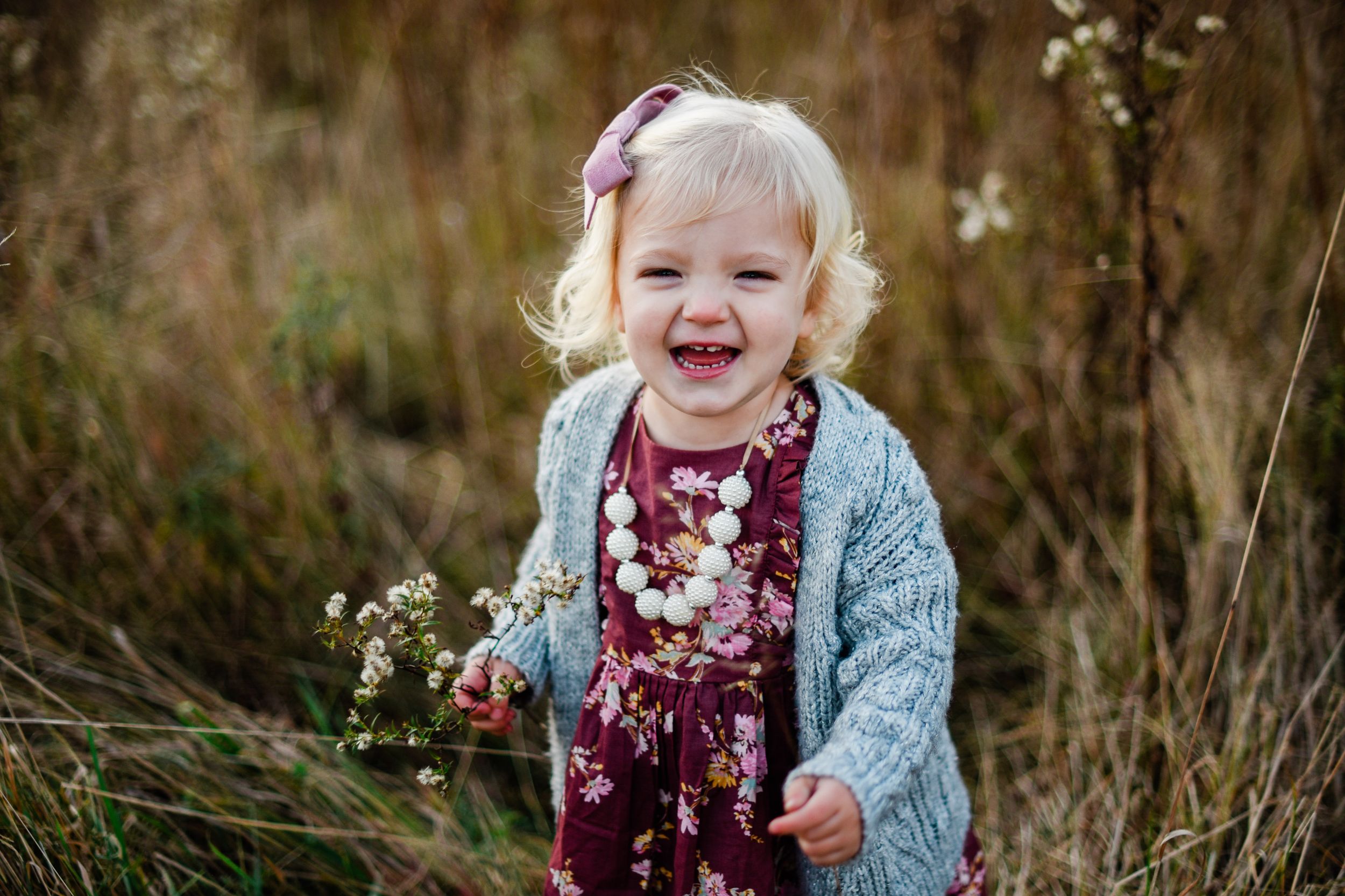 Young child laughing outdoors among autumn wildflowers wearing a floral dress.