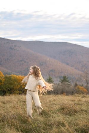 A person in white clothing dancing and twirling in a mountain meadow with autumn foliage in the background.