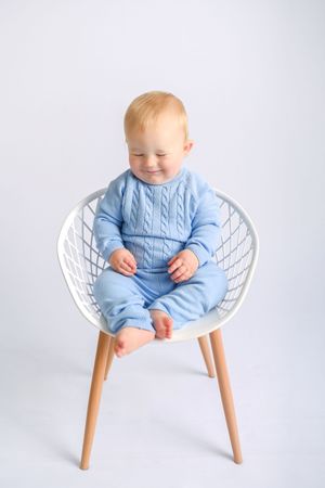 Baby wearing light blue cable knit outfit sits in modern white wire chair.