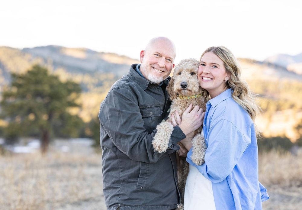 A couple stands outdoors in a mountain landscape while holding their small brown dog on a sunny afternoon.