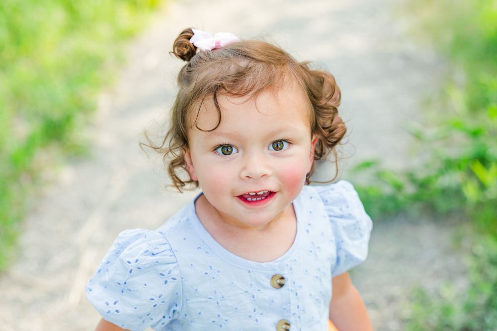 Adorable toddler girl with curly hair and pink bow smiling outdoors in a light blue dress on a sunny day.