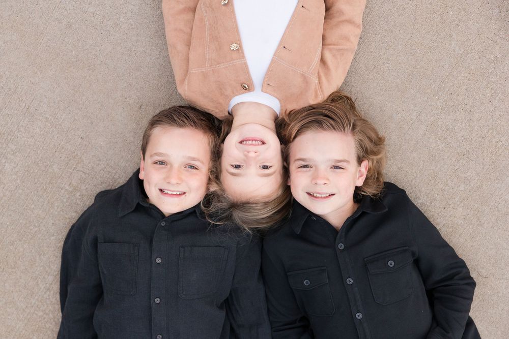 Three smiling children lying on the ground looking up, two boys in black shirts and a girl in a peach cardigan.