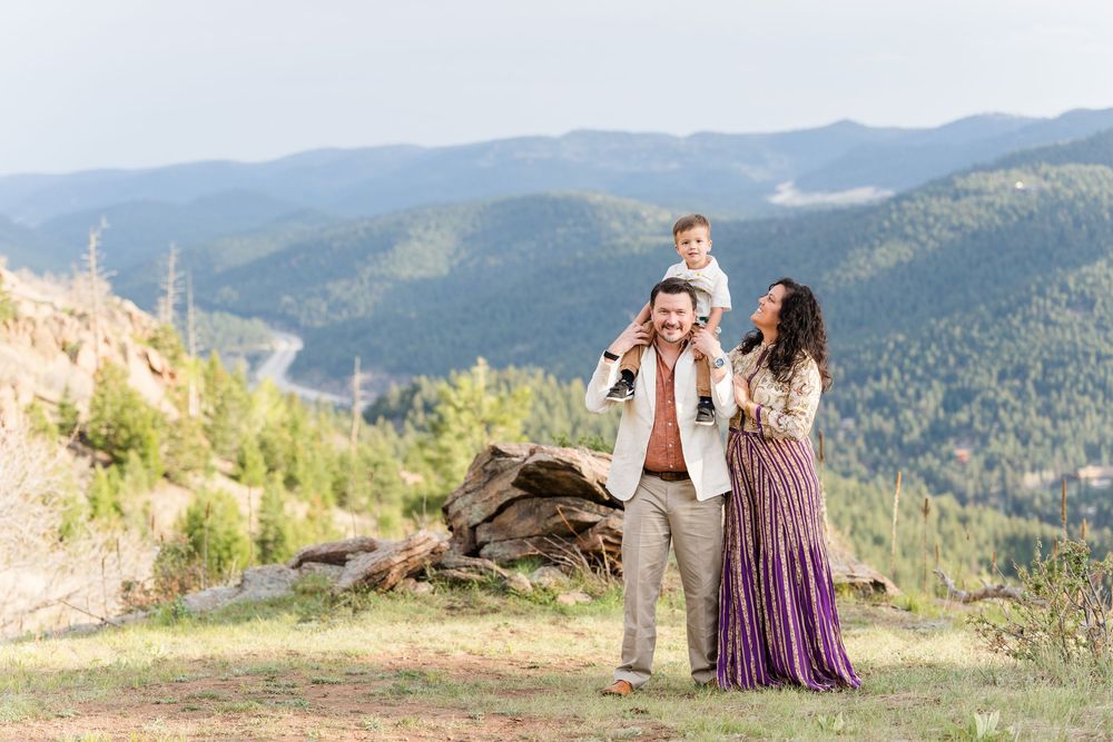 Family posing on mountain overlook, father carrying son on shoulders, mother in purple dress, scenic forest valley backdrop.