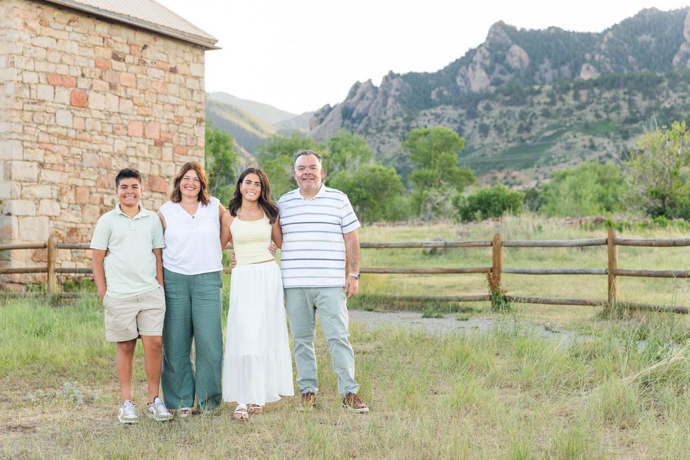 Family of four smiling outdoors near a rustic stone building with mountain views and a wooden fence.