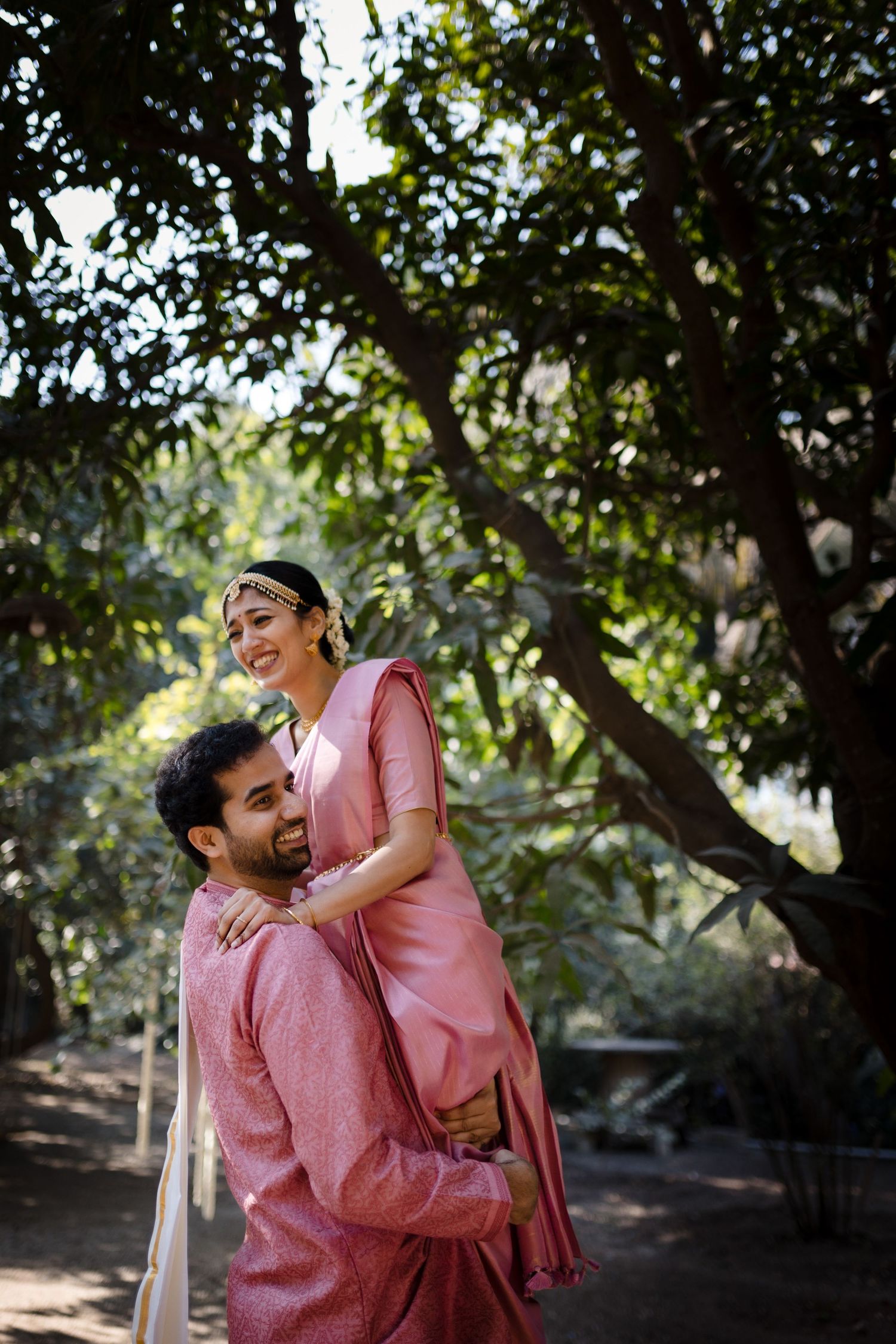 A couple wearing matching pink outfits share a joyful moment under trees.