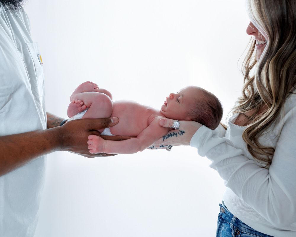 Newborn baby being gently held by adults in white shirts against soft background.
