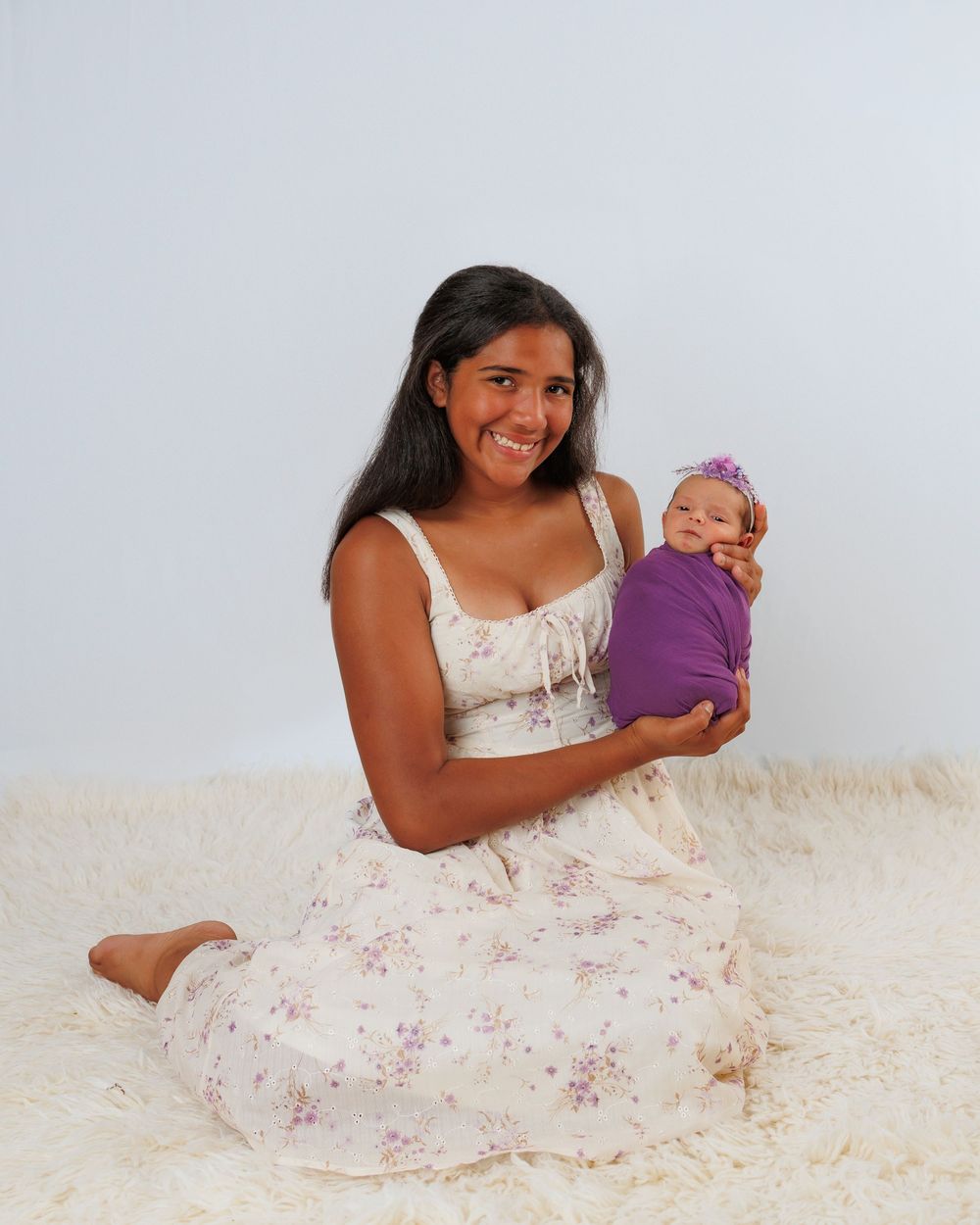 A person in a floral dress sits on a cream carpet while holding a small baby doll wrapped in purple fabric.