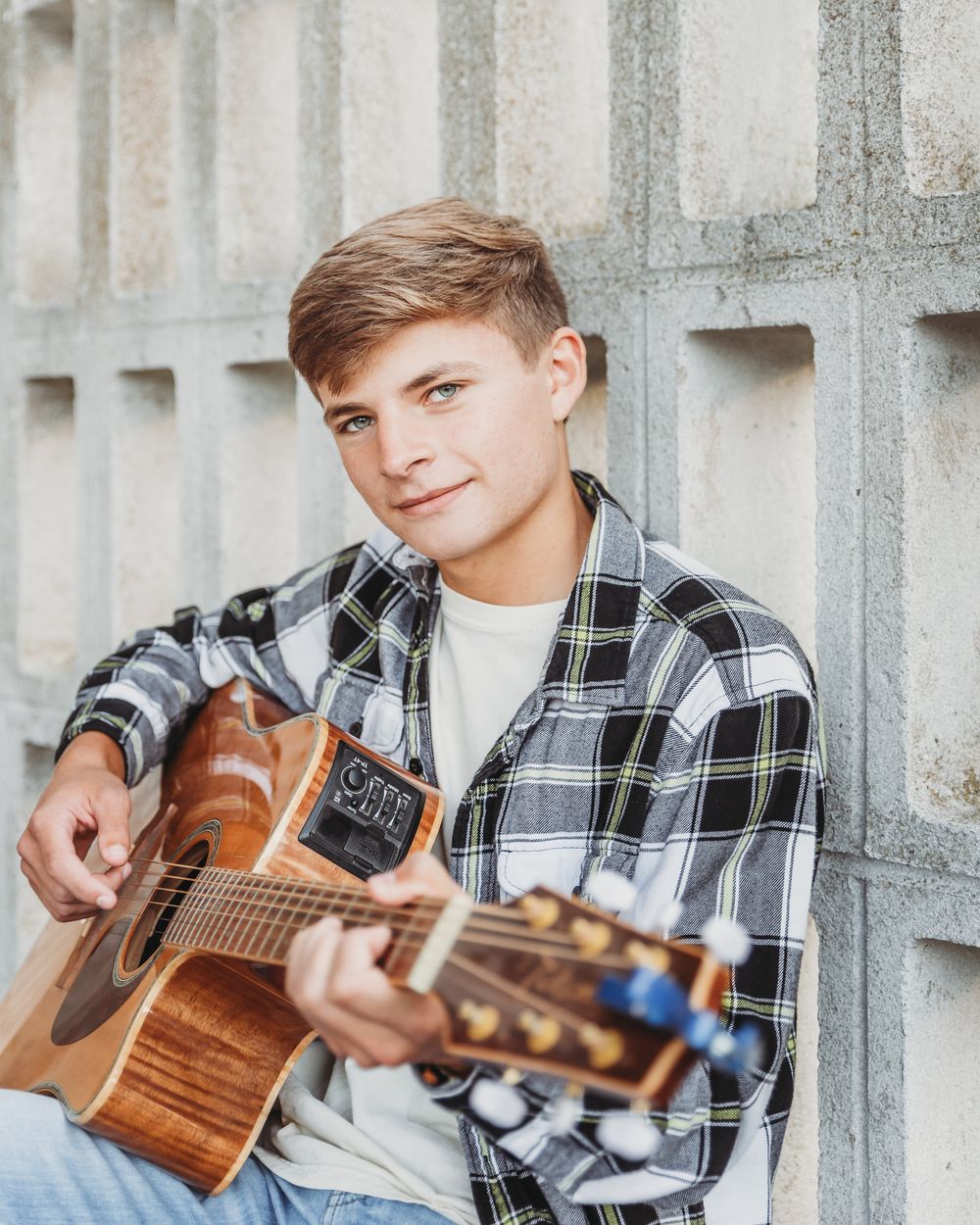 Young musician in plaid flannel shirt plays acoustic guitar while sitting against concrete wall.