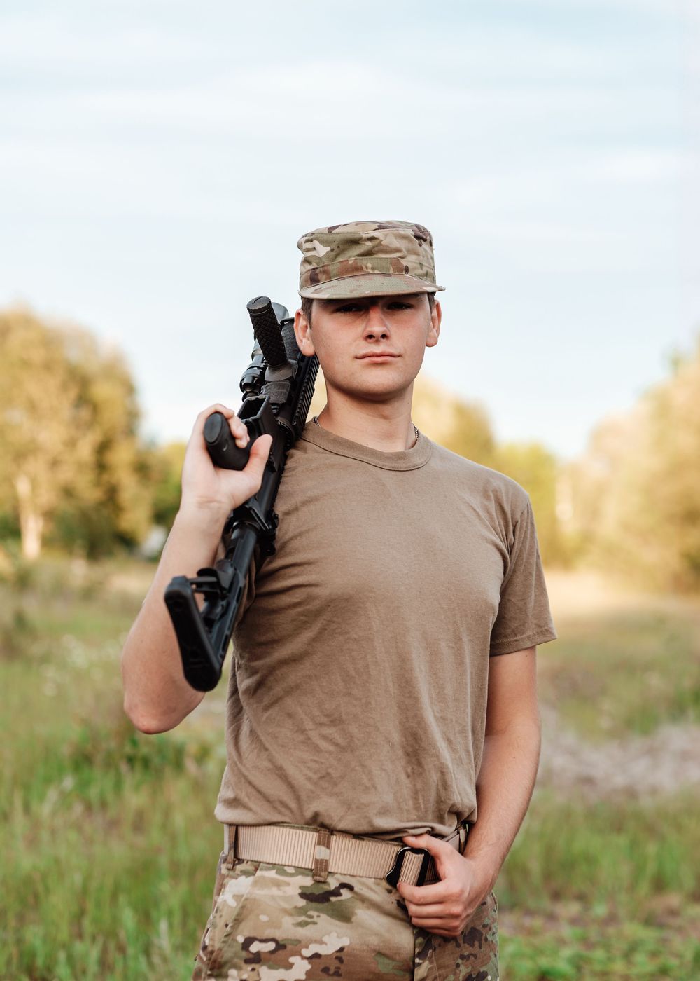 Military service member in combat uniform and cap holding a rifle while standing in an outdoor field setting.