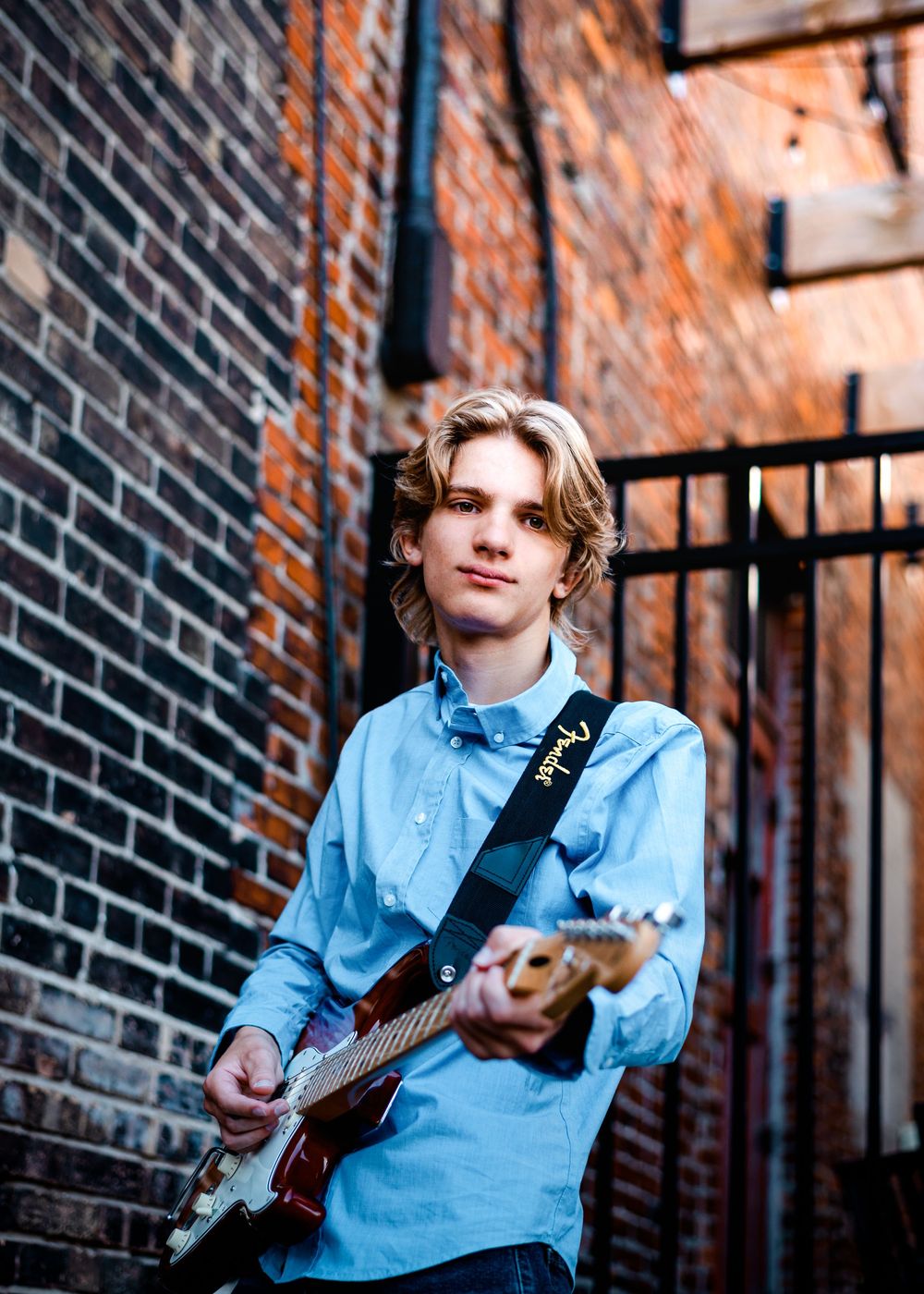 Musician in a light blue button-down shirt plays bass guitar while posing against a rustic brick wall outdoors.
