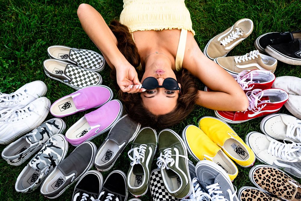 Someone lying on grass surrounded by a colorful collection of sneakers and canvas shoes arranged in a circular pattern.