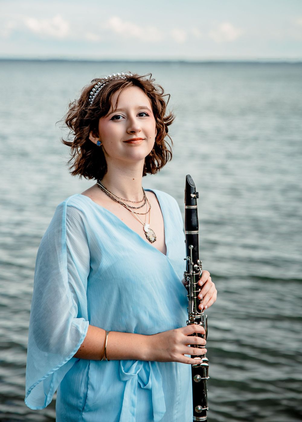Musician posing in light blue dress with clarinet against ocean backdrop on overcast day.