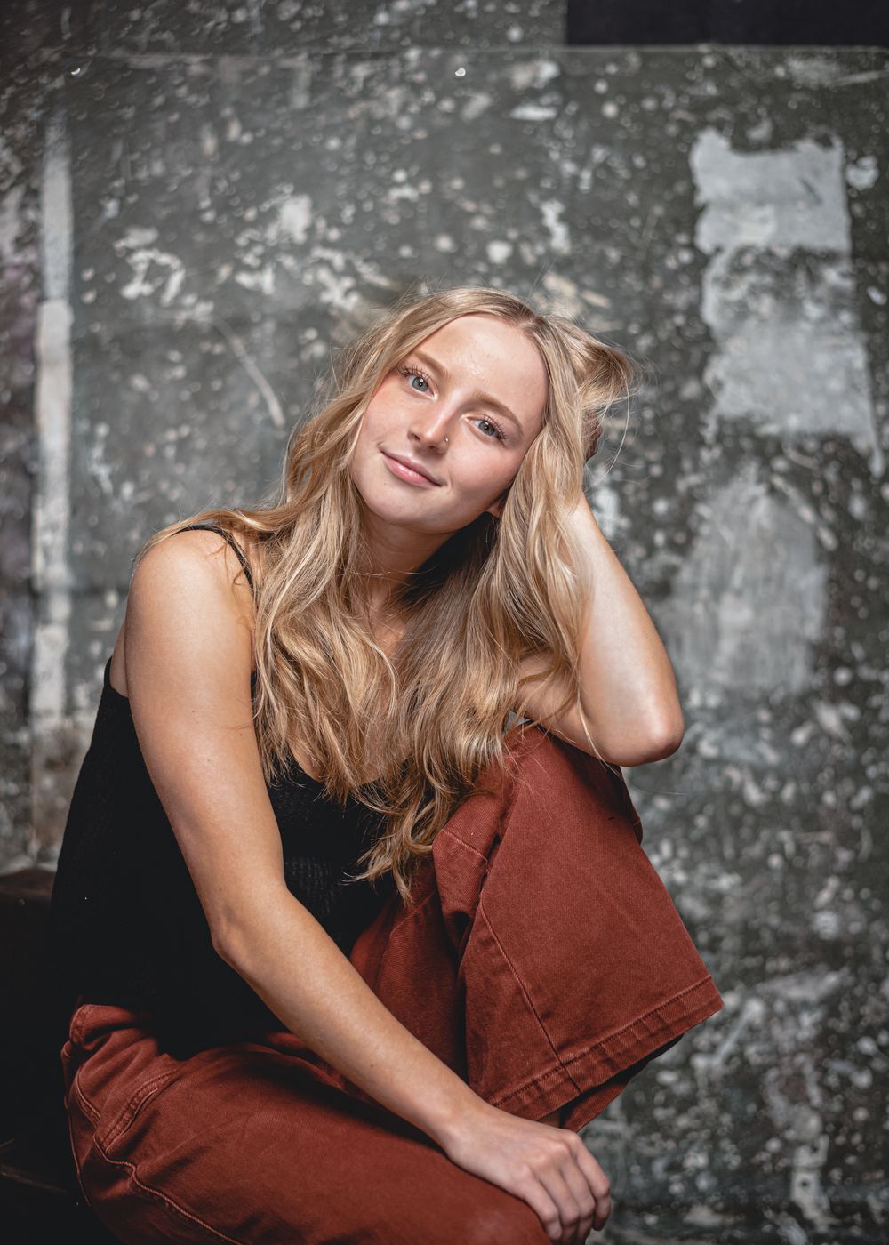 Casual portrait with blonde hair and burnt orange pants against textured concrete wall in moody lighting.