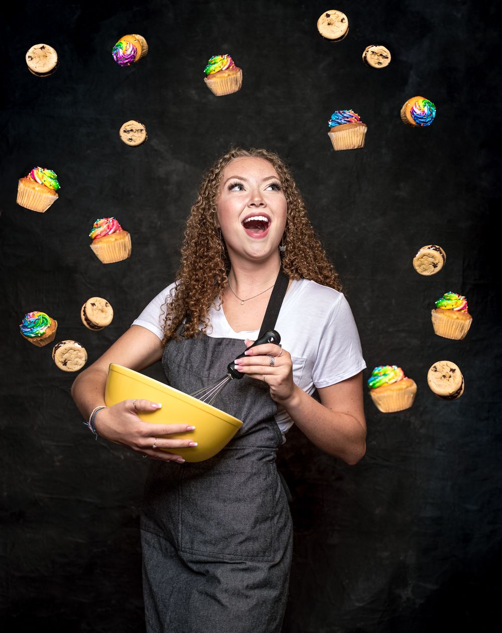 A baker in a gray apron with a mixing bowl joyfully laughs while colorful cupcakes float around on a black background.