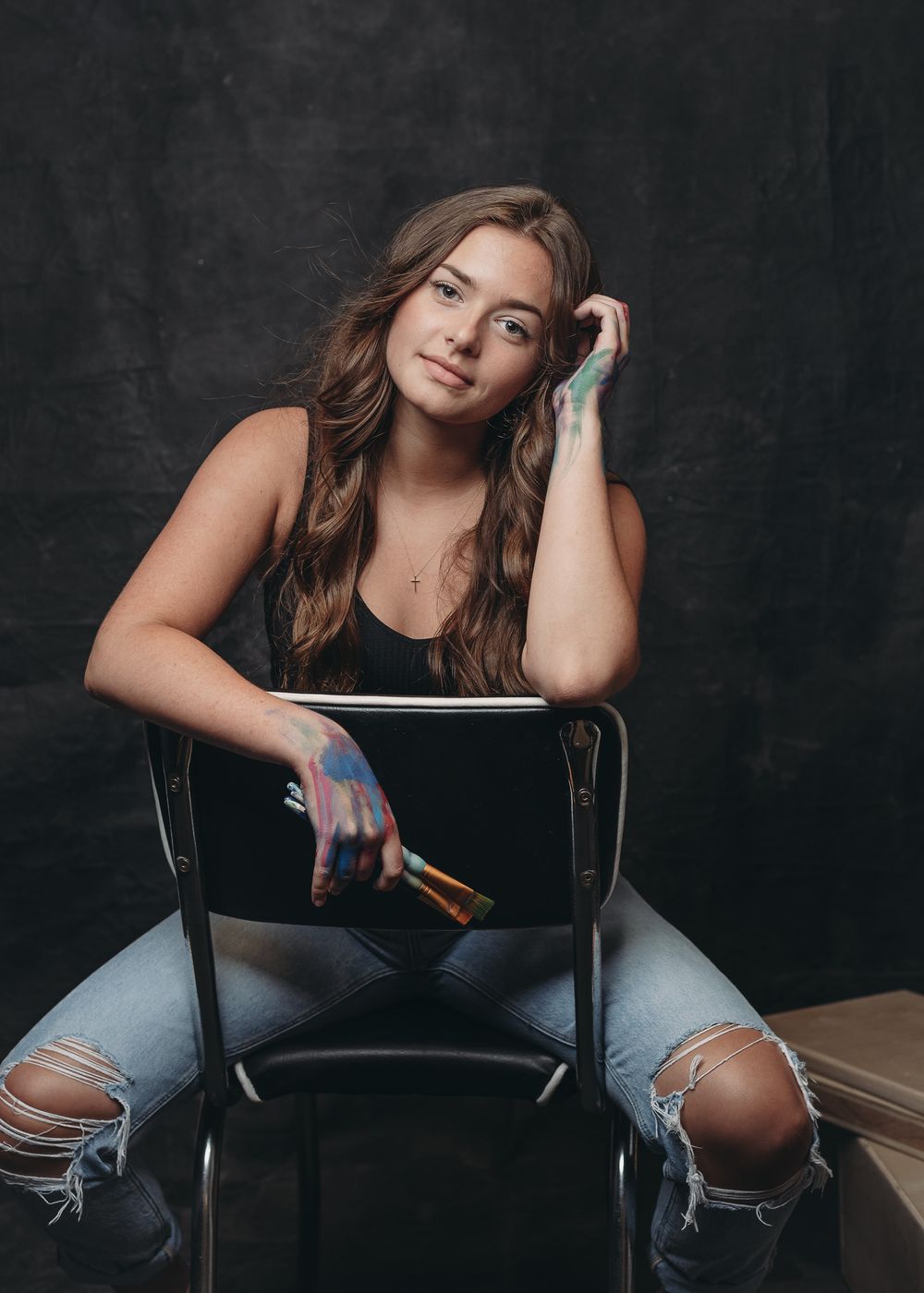 Artistic portrait of someone in a black tank top and ripped jeans sitting casually on a chair against a dark background.