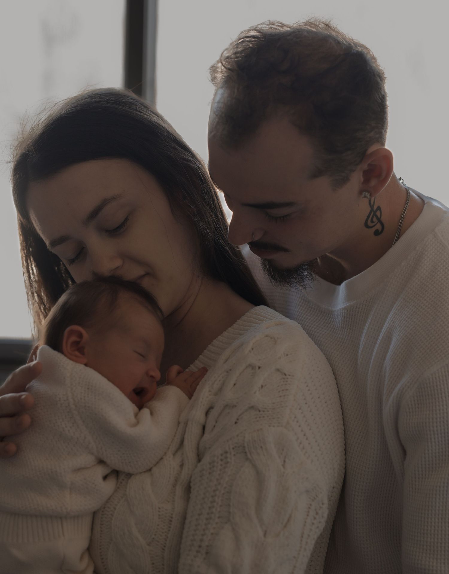 A tender family moment as parents in white sweaters embrace their sleeping baby in a cozy indoor setting.