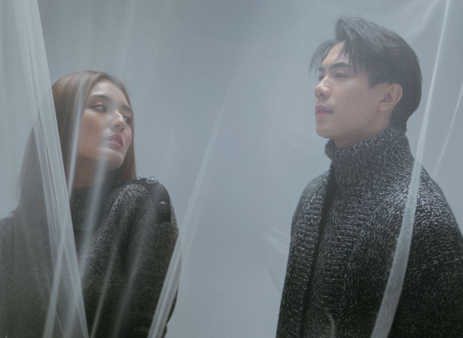 Two people in dark sweaters stand behind translucent curtains creating a dreamy, ethereal mood in soft lighting.