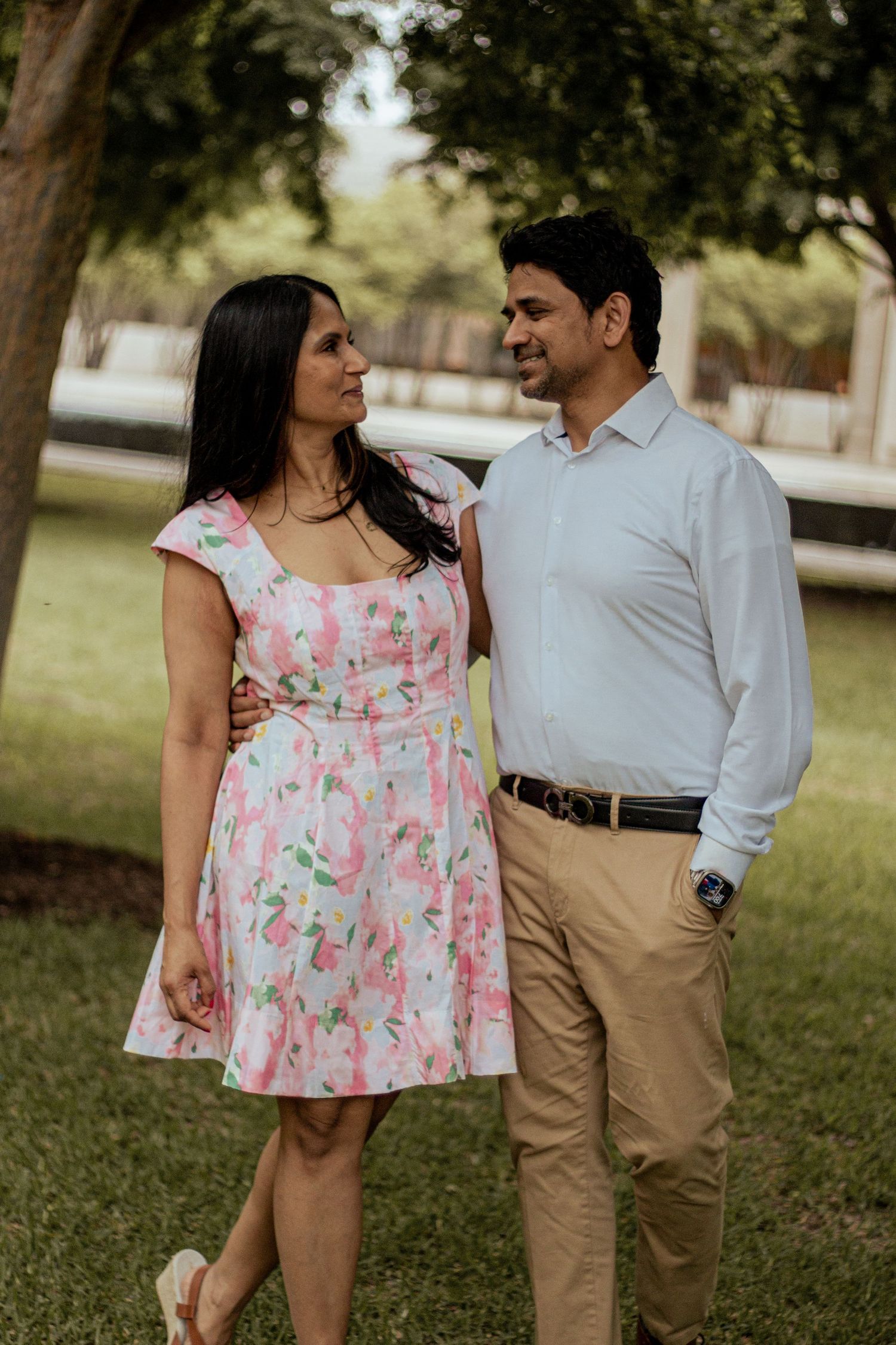 A couple stands lovingly in a park setting, she wearing a pink floral dress and he in a white shirt and khakis.