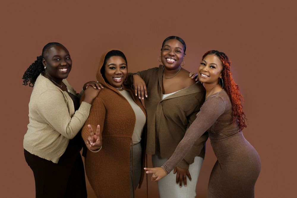 Four friends laughing together while posing against brown background wearing neutral-toned outfits.